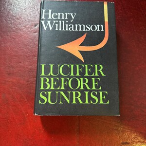 May include: A hardcover book titled Lucifer Before Sunrise by Henry Williamson. The book cover is black with white text for the author's name, bright green text for the title, and a stylized orange arrow graphic pointing toward the left side.