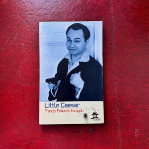 May include: A paperback book titled Little Caesar by Francis Edwards Faragoh. The cover features a blue-toned black and white photograph of a man in a dark suit holding a small handgun. The bottom section is cream-colored with the title and author.
