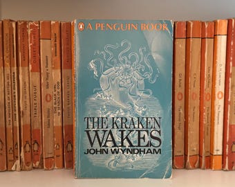 May include: A vintage Penguin Book edition of "The Kraken Wakes" by John Wyndham, with a teal cover and kraken design. It's surrounded by other Penguin paperbacks with orange spines, a classic book collection.