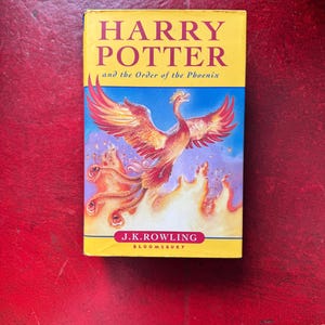 May include: A hardback book titled Harry Potter and the Order of the Phoenix by J.K. Rowling. The yellow and blue cover features a large, vibrant orange and red phoenix rising from flames, with the author's name printed in a red oval at the bottom.