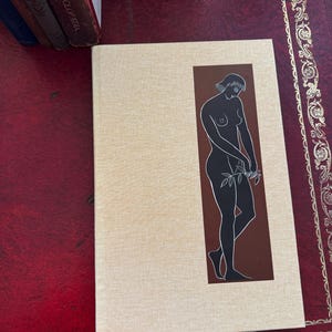 May include: A hardcover book with a beige cover and a brown rectangular graphic featuring a black silhouette of a person holding leaves. The book is on a red surface with gold decorative trim. Several other books are visible in the background.