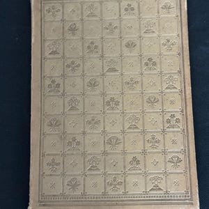 Puede incluir: Una cubierta de libro vintage con un patrón en relieve detallado. La cubierta es de color beige claro, con una cuadrícula de cuadrados llenos de diseños florales y geométricos. Los bordes de la cubierta muestran signos de desgaste.