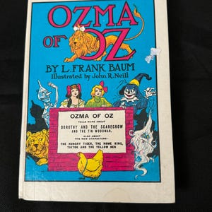 May include: A vintage book cover for "Ozma of Oz" by L. Frank Baum, illustrated by John R. Neill. The cover features a colorful illustration of characters from the book, including Dorothy, the Scarecrow, the Tin Woodman, and Ozma.