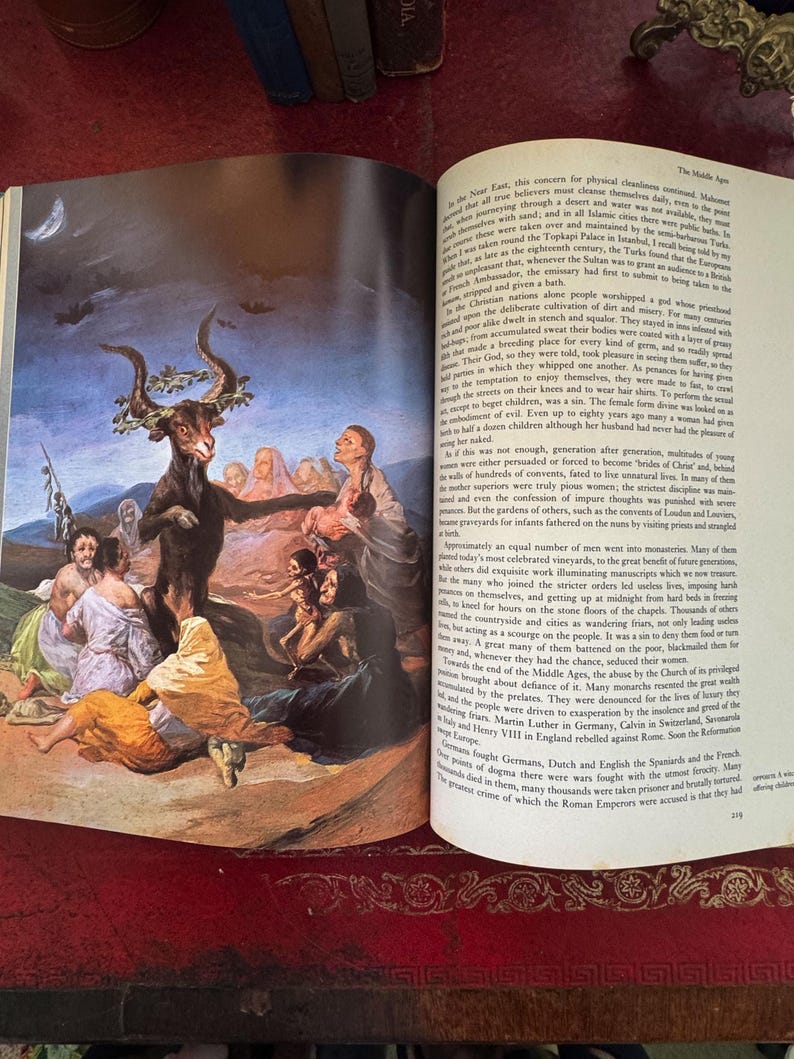 May include: An open book with a detailed illustration depicting a goat-like figure with horns surrounded by people in a historical setting. The page also contains printed text.