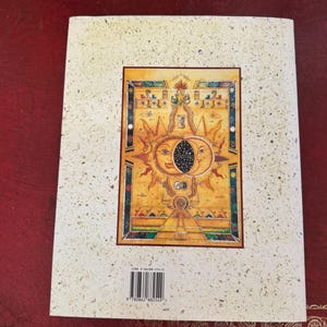 May include: A book cover with a textured, off-white background. The cover features a central rectangular image with a sun and moon design in gold, orange, and blue. The image has intricate details and a border. The ISBN barcode is visible.