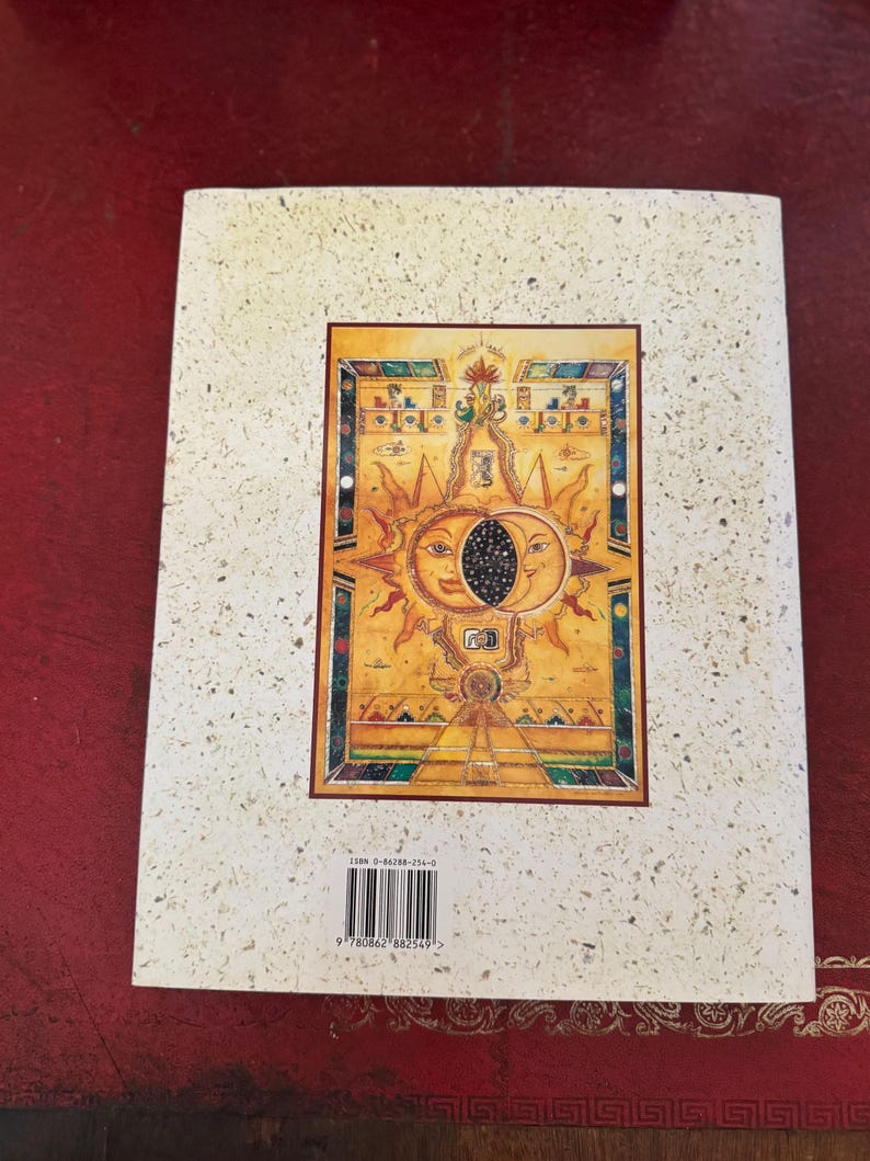 May include: A book cover with a textured, off-white background. The cover features a central rectangular image with a sun and moon design in gold, orange, and blue. The image has intricate details and a border. The ISBN barcode is visible.