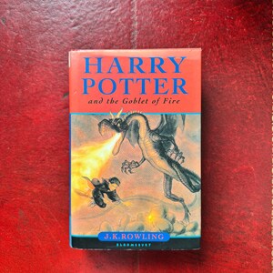 May include: A hardback book titled Harry Potter and the Goblet of Fire by J.K. Rowling, published by Bloomsbury. The cover features a blue title, a black dragon breathing fire, and a figure on a broomstick against a light background.