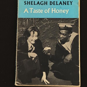 Op de afbeelding: Vintage paperback boekomslag voor "A Taste of Honey" van Shelagh Delaney. De omslag toont een zwart-witfoto van twee mensen, een in een matrozenuniform en de ander in een donker pak, met de titel en de naam van de auteur in blauw.