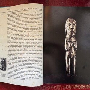 May include: A silver-toned, stylized figurine of a person with a long head and hands clasped in front. The figure is on a black background, with a book page visible on the left.