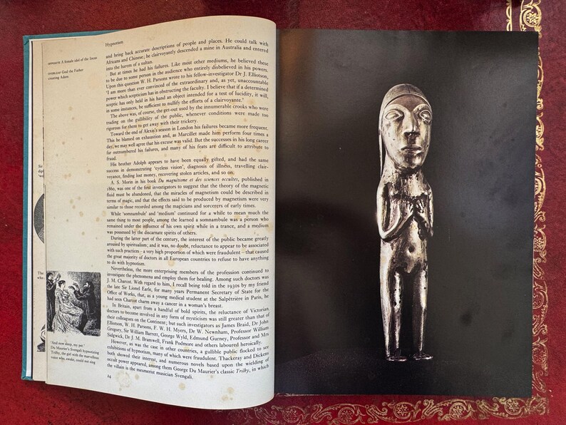 May include: A silver-toned, stylized figurine of a person with a long head and hands clasped in front. The figure is on a black background, with a book page visible on the left.