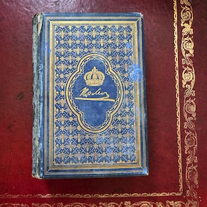 May include: A blue hardback book with gold-embossed bee patterns and a central emblem featuring a crown above the handwritten text Napoléon. The book cover has a decorative gold border and shows signs of wear along the spine edge.