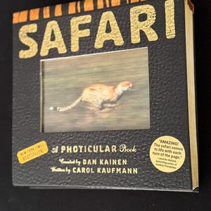 Peut inclure: Un livre cartonné intitulé "SAFARI" avec des lettres dorées et une fenêtre rectangulaire montrant un animal. Le livre est un "PHOTICULAR Book" de Dan Kainen et Carol Kaufmann. Un autocollant "NEW YORK TIMES BESTSELLER" est à gauche.