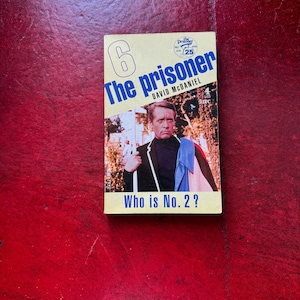 May include: A paperback book titled The Prisoner by David McDaniel, featuring a large number 6, a 25th-anniversary logo, and a photograph of a man in a black blazer with a blue scarf. The cover text includes Who is No. 2? and the number 4.