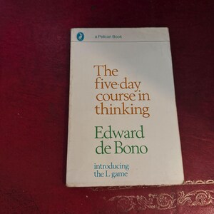 May include: A white paperback book titled The five-day course in thinking by Edward de Bono, featuring the subtitle introducing the L game. The cover includes a small blue pelican logo and the text a Pelican Book in a clean, minimalist layout.