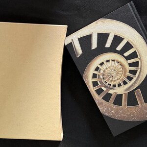 Könnte beinhalten: Eine goldfarbene Schachtel und ein Buch mit schwarzem Einband. Das Buch zeigt ein Spiralmuster in Gold- und Brauntönen. Die Schachtel ist rechteckig und hat eine glatte, matte Oberfläche.