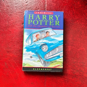 May include: A hardcover book titled Harry Potter and the Chamber of Secrets by J.K. Rowling, published by Bloomsbury. The cover features a blue flying car with two people and an owl inside, set against a blue sky and green landscape background.