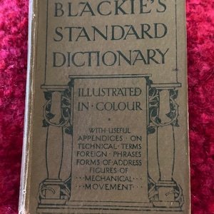 May include: A vintage book titled "Blackie's Standard Dictionary" with the words "Illustrated in Colour" prominently displayed. The cover is a muted brown with decorative elements and text. The book's spine is visible, and the background is a textured, reddish-pink.