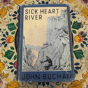 Pode incluir: Um livro vintage intitulado "Sick Heart River" de John Buchan. A capa apresenta uma ilustração a preto e branco de uma cena de montanha com uma figura. O livro tem lombada e borda azuis, sobre uma superfície com padrões florais.