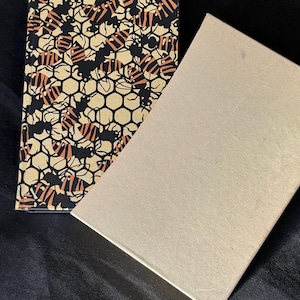 Könnte beinhalten: Eine rechteckige Schachtel mit Deckel, neben einem Buch. Das Buchcover zeigt ein Muster aus Bienen und Waben in Schwarz, Beige und Orange. Der Deckel der Schachtel ist einfarbig hellbeige.
