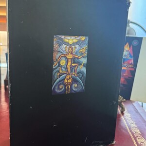 May include: A black book with a colorful illustration of a figure with outstretched arms, a sun, and other figures. The book is standing upright on a red surface.