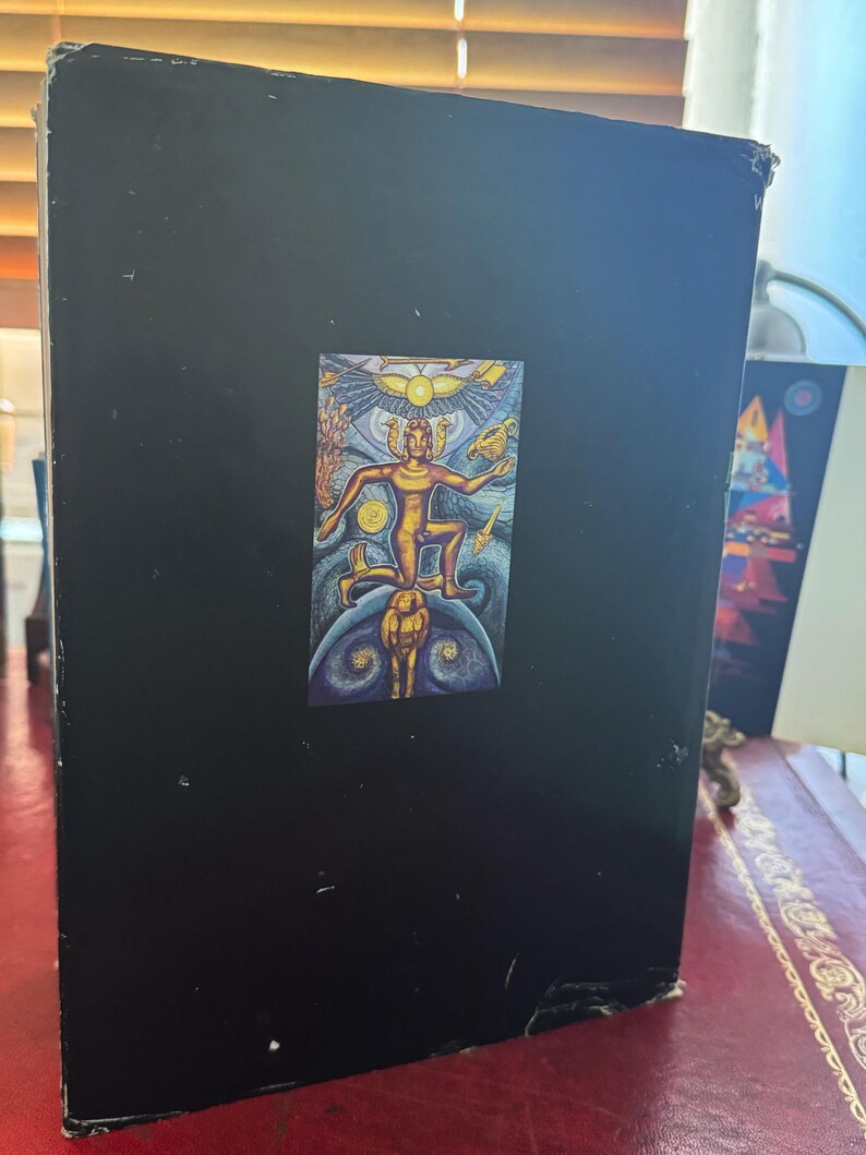 May include: A black book with a colorful illustration of a figure with outstretched arms, a sun, and other figures. The book is standing upright on a red surface.