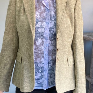 May include: A beige blazer with a light blue floral patterned shirt underneath. The blazer has a textured fabric and a single button closure.