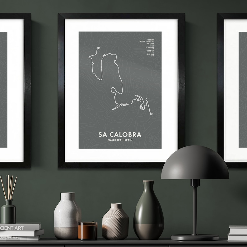 May include: A framed print of a topographic map of the Sa Calobra cycling route in Mallorca, Spain. The print is in a black frame and is hanging on a dark green wall. There are decorative items on a shelf below the print, including a lamp, vases, and books.