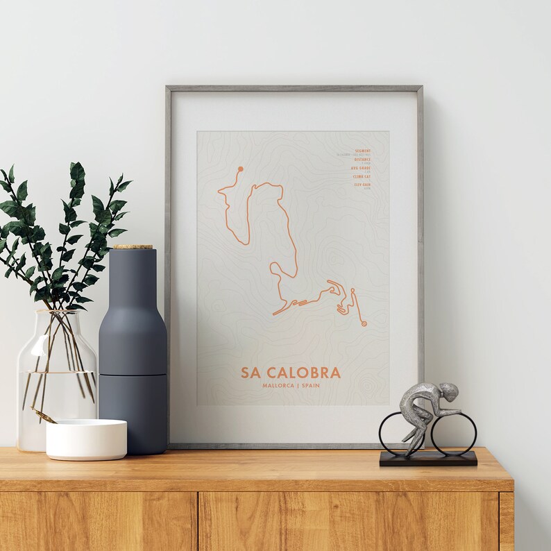May include: A framed print of a topographic map of the Sa Calobra cycling route in Mallorca, Spain. The print is on a white background with a brown frame. A silver figurine of a cyclist is on a black base in front of the print.