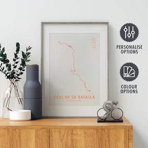 May include: A framed print of a cycling route map with the text "COLL DE SA BATALLA, MALLORCA, SPAIN". The print is on a wooden shelf with a grey bottle and a silver bicycle figurine.