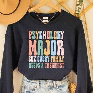 May include: A navy blue cropped sweatshirt with the text "Psychology Major Cuz Every Family Needs A Therapist" in rainbow colours. The sweatshirt is worn with denim shorts and a straw bag.