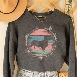 May include: Dark heather grey sweatshirt with a vintage-style graphic of a dachshund in a pink, blue, and green circle. The sweatshirt is shown on a model wearing denim shorts.
