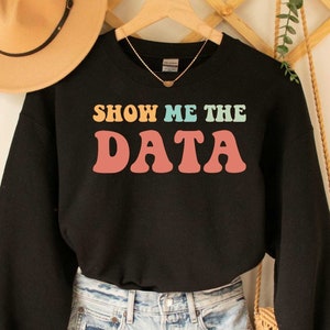May include: Black sweatshirt with the text "Show Me The Data" in rainbow colors. The sweatshirt is being modeled with a pair of light blue jeans and a straw bag.