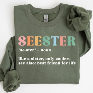 May include: A military green sweatshirt with the word "SEESTER" in colorful letters. Below the word is a definition: "like a sister, only cooler, see also: best friend for life."