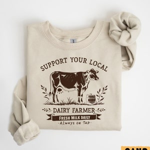 May include: A beige sweatshirt with a brown graphic of a cow and the text "Support Your Local Dairy Farmer - Fresh Milk Daily - Always on Tap."