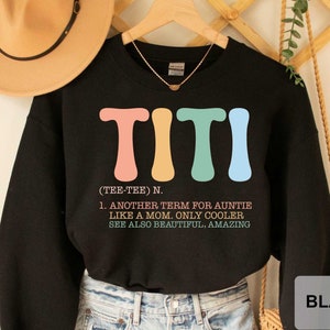 May include: Black sweatshirt with a colorful graphic that says "TITI (TEE-TEE) N. 1. ANOTHER TERM FOR AUNTIE LIKE A MOM. ONLY COOLER SEE ALSO BEAUTIFUL, AMAZING"