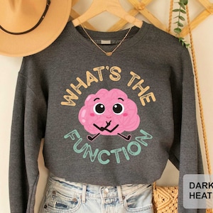 May include: Dark heather grey sweatshirt with a pink brain cartoon graphic and the text "What's the Function".