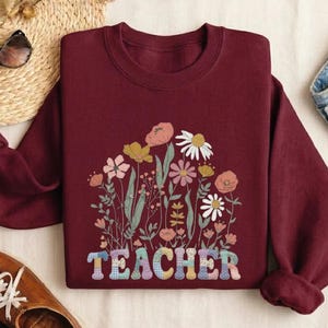 May include: Maroon sweatshirt with a floral design and the word "TEACHER" in colorful letters. The sweatshirt features a crew neck and long sleeves. The floral design includes various flowers in shades of pink, yellow, and white.