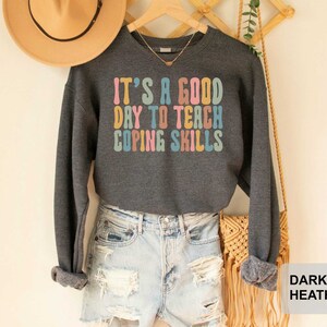 May include: Dark heather gray sweatshirt with the text "IT'S A GOOD DAY TO TEACH COPING SKILLS" in colorful retro font. The long-sleeved top is paired with light-wash denim shorts. A tan hat hangs in the background.
