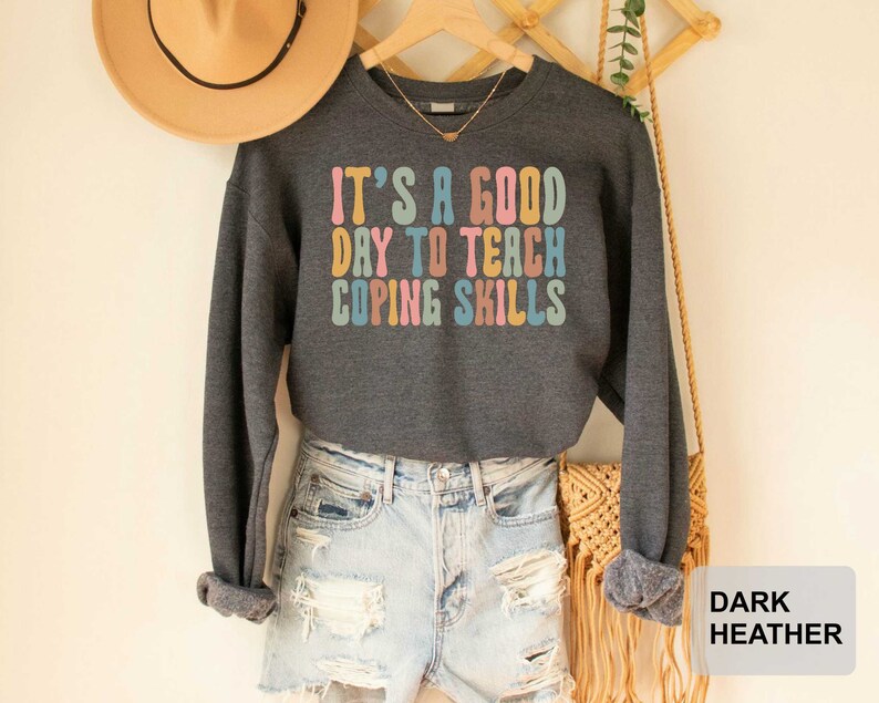 May include: Dark heather gray sweatshirt with the text "IT'S A GOOD DAY TO TEACH COPING SKILLS" in colorful retro font. The long-sleeved top is paired with light-wash denim shorts. A tan hat hangs in the background.