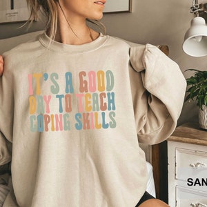 May include: Sand-colored sweatshirt with the text "IT'S A GOOD DAY TO TEACH COPING SKILLS" in colorful, retro-style lettering. The sweatshirt has a crew neck and long sleeves, and is made of a soft material.