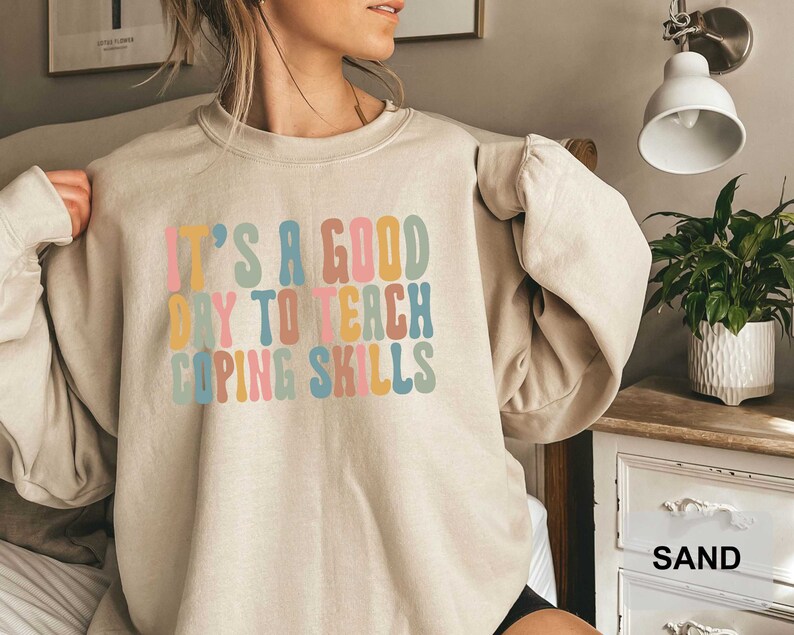 May include: Sand-colored sweatshirt with the text "IT'S A GOOD DAY TO TEACH COPING SKILLS" in colorful, retro-style lettering. The sweatshirt has a crew neck and long sleeves, and is made of a soft material.
