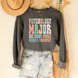 May include: A dark grey sweatshirt with the text "Psychology Major Cuz Every Family Needs A Therapist" in colourful letters. The sweatshirt is being worn by a person wearing denim shorts and a straw hat.