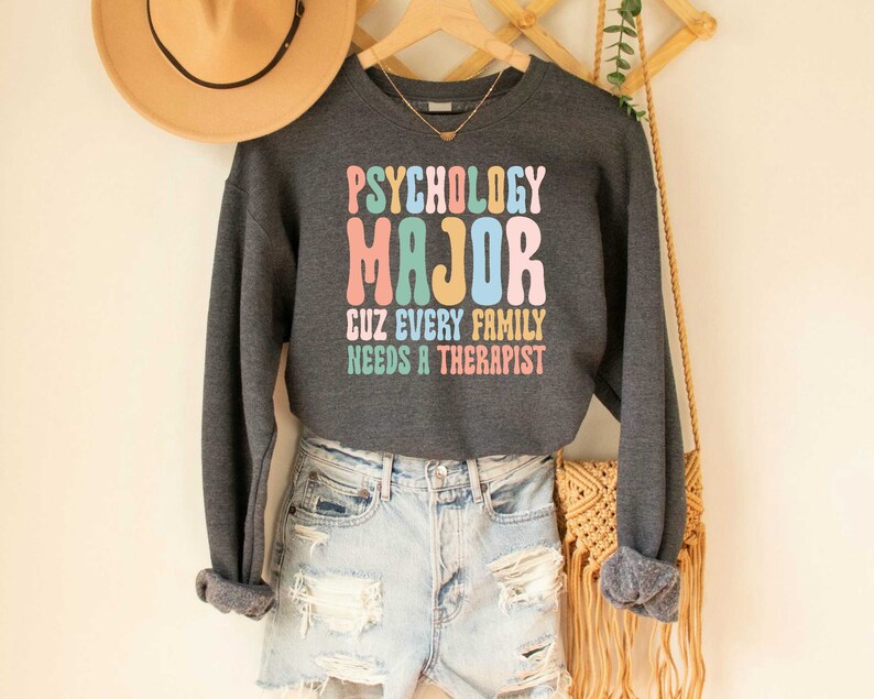 May include: A dark grey sweatshirt with the text "Psychology Major Cuz Every Family Needs A Therapist" in colourful letters. The sweatshirt is being worn by a person wearing denim shorts and a straw hat.
