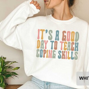 May include: White sweatshirt with the text "IT'S A GOOD DAY TO TEACH COPING SKILLS" in colorful, retro-style lettering. The long-sleeved top is a casual, comfortable style.