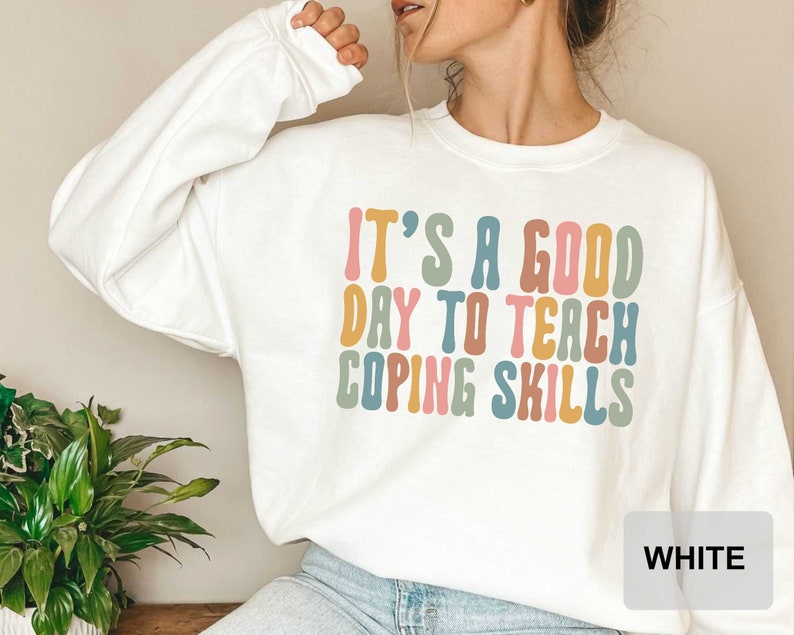 May include: White sweatshirt with the text "IT'S A GOOD DAY TO TEACH COPING SKILLS" in colorful, retro-style lettering. The long-sleeved top is a casual, comfortable style.