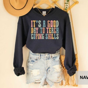May include: Navy blue sweatshirt with the text "IT'S A GOOD DAY TO TEACH COPING SKILLS" in colorful retro font. The long-sleeved top is displayed on a hanger with a straw hat and a necklace.