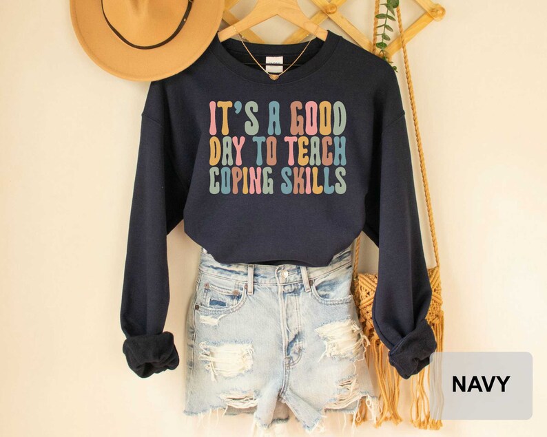 May include: Navy blue sweatshirt with the text "IT'S A GOOD DAY TO TEACH COPING SKILLS" in colorful retro font. The long-sleeved top is displayed on a hanger with a straw hat and a necklace.