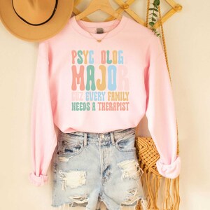 May include: Pink sweatshirt with the text "PSYCHOLOG MAJOR CUZ EVERY FAMILY NEEDS A THERAPIST" in rainbow colours.