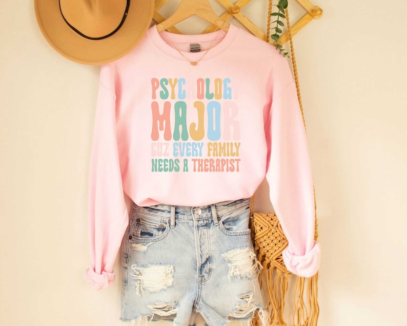 May include: Pink sweatshirt with the text "PSYCHOLOG MAJOR CUZ EVERY FAMILY NEEDS A THERAPIST" in rainbow colours.