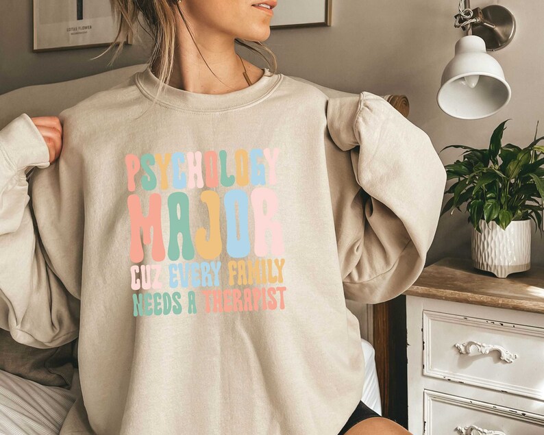 May include: A beige sweatshirt with the text "Psychology Major Cuz Every Family Needs A Therapist" in colourful, retro-style lettering.